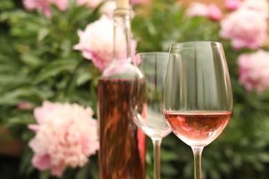 Glasses and bottle with rose wine against beautiful peonies, closeup. Space for text