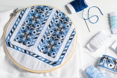 Shirt with blue embroidery design in hoop, needle and threads on white wooden table, flat lay. National Ukrainian clothes
