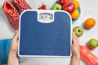 Woman holding scales over table with healthy food and sport equipment, top view. Weight loss