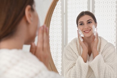 Young woman applying cleansing foam onto her face near mirror in bathroom
