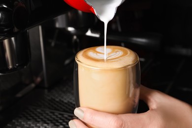 Photo of Barista pouring steamed milk from pitcher into glass cup of fresh aromatic coffee in cafe, closeup
