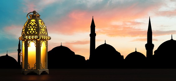 Decorative Arabic lantern on wooden surface and silhouette of mosque at sunset on background, banner design