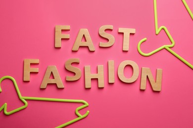 Photo of Phrase FAST FASHION made of wooden letters and bright hangers on pink background, flat lay