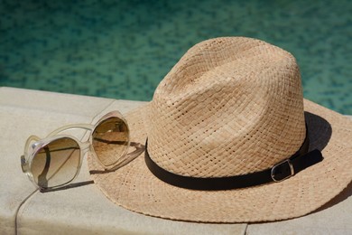 Stylish hat and sunglasses near outdoor swimming pool on sunny day, closeup. Beach accessories