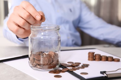 Man putting coin into glass jar at table indoors, closeup
