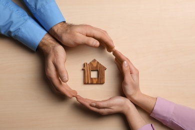 Couple holding hands near figure of house on wooden background, top view. Home insurance