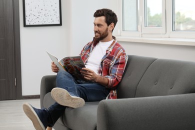 Smiling bearded man reading magazine on sofa at home