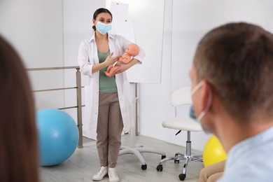 Doctor in protective mask teaching pregnant woman and her husband to take care of baby indoors