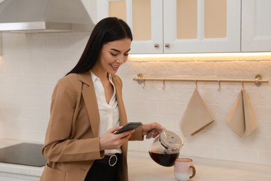 Photo of Young woman with smartphone pouring coffee into cup at home. Morning routine