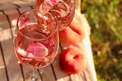 Photo of Glasses of delicious rose wine with petals and peaches on white picnic blanket outside. Space for text