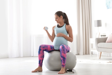 Young pregnant woman in fitness clothes lifting dumbbell at home