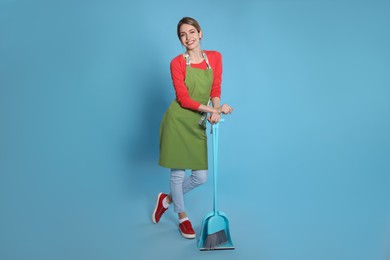 Young housewife with broom and dustpan on light blue background