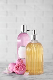 Stylish dispenser with liquid soap, bottle of shower gel and flowers on white marble table