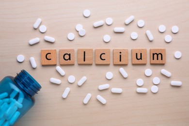 Word Calcium made of cubes with letters, medical bottle and pills on wooden table, flat lay