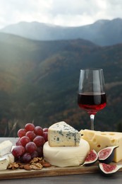 Different types of delicious cheeses, fruits and wine on wooden table against mountain landscape