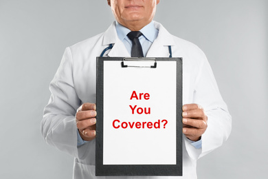Doctor holding clipboard with text ARE YOU COVERED? on light background, closeup. Insurance concept