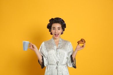 Excited young woman in silk bathrobe with hair curlers having breakfast on orange background