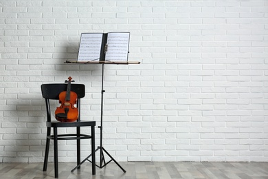 Violin, chair and note stand with music sheets near brick wall. Space for text