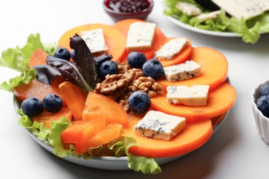 Photo of Delicious persimmon with blue cheese, blueberries, lettuce and walnuts on white table, closeup