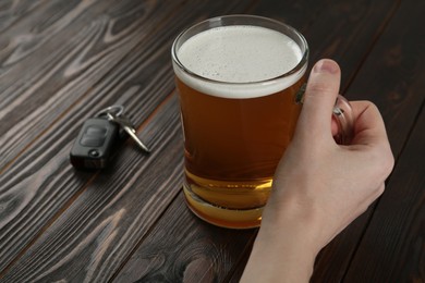 Man holding glass of alcoholic beer near car key at wooden table, closeup. Dangerous drinking and driving