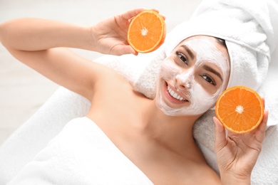 Pretty woman with rejuvenating facial mask holding sliced orange in spa salon, above view