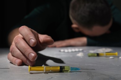 Photo of Overdosed man at grey table, focus on syringe with drugs
