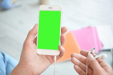 Image of Chroma key compositing. Woman holding smartphone with green screen and earphones indoors, closeup. Mockup for design