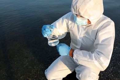 Scientist in chemical protective suit with conical flask taking sample from river for analysis