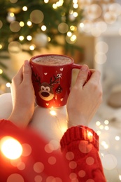 Woman holding festive cup with coffee near Christmas tree indoors, closeup