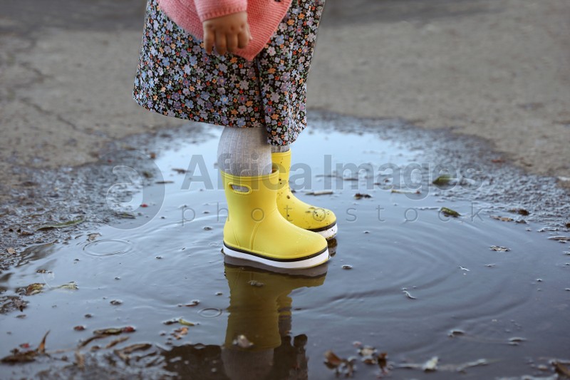 Photo of Little girl wearing rubber boots standing in puddle outdoors, closeup. Autumn walk