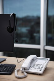 Stationary phone near modern computer with headset on wooden desk indoors. Hotline service