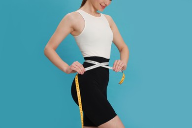 Happy woman in sportswear measuring waist with tape on light blue background, closeup