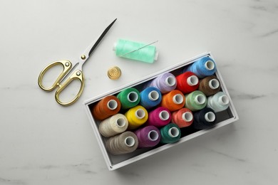 Box with colorful sewing threads, scissors and thimble on white marble table, flat lay