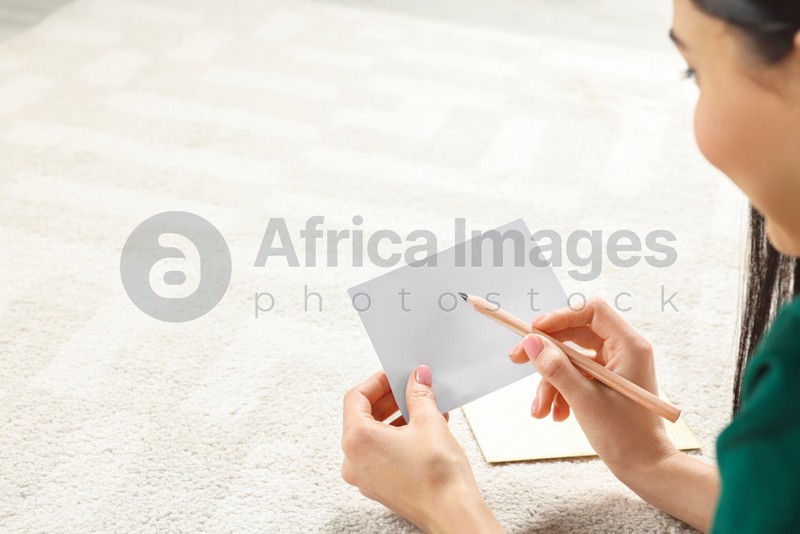 Photo of Woman writing message in greeting card on carpet in room, closeup. Space for text