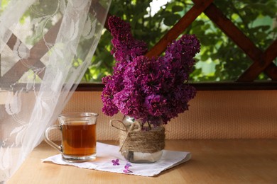 Photo of Bouquet with beautiful lilac flowers and glass cup of tea on wooden table indoors