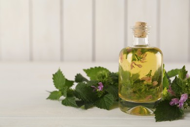 Glass bottle of nettle oil with flowers and leaves on white wooden table, closeup. Space for text