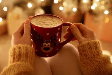 Woman with cup of drink and blurred Christmas lights on background, closeup
