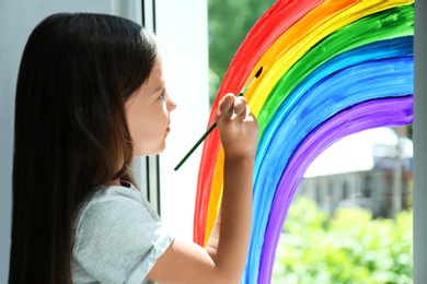 Little girl drawing rainbow on window indoors. Stay at home concept