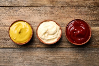 Ketchup, mustard and mayonnaise in bowls on wooden table, flat lay
