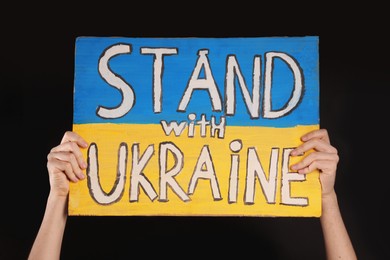 Teenage boy holding poster Stand with Ukraine against black background, closeup