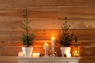 Small potted firs, candles and decor elements on white mantel near wooden wall
