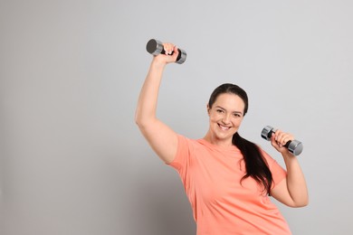 Happy overweight woman doing exercise with dumbbells on grey background, space for text