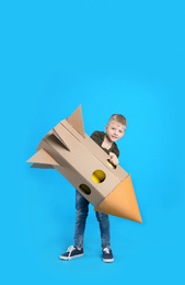 Little child playing with rocket made of cardboard box on light blue background. Space for text