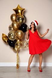 Happy woman in Santa hat with champagne and air balloons near beige wall indoors. Christmas party