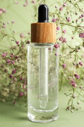 Bottle of face serum and beautiful flowers on light green background, closeup