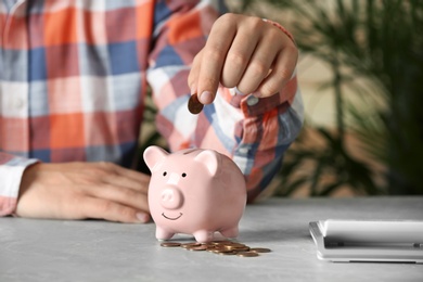 Man putting coin into piggy bank at grey marble table, closeup. Money savings