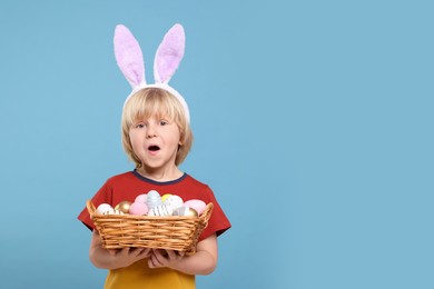 Photo of Happy boy in bunny ears headband holding wicker basket with painted Easter eggs on turquoise background. Space for text