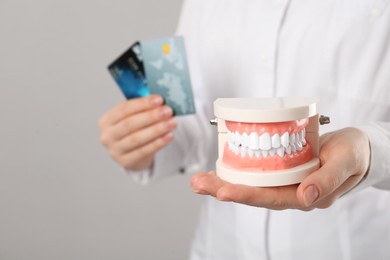Dentist holding educational typodont model and credit cards on grey background, closeup. Expensive treatment