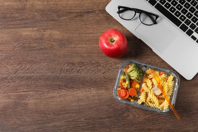 Photo of Container of tasty food, laptop, apple and glasses on wooden table, flat lay with space for text. Business lunch