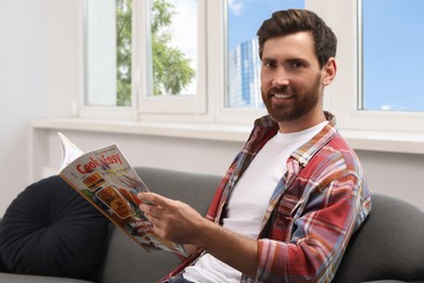 Smiling bearded man with magazine on sofa indoors